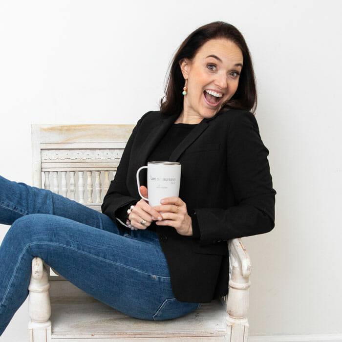 Woman with excited and happy expression on face wearing black blazer and black shirt with blue jeans sitting on chair holding cup with The Game On Girlfriend Podcast